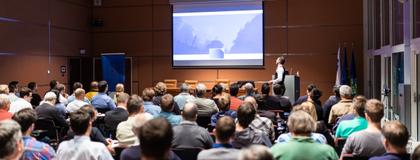 A speaker stands in front of a crowded lecture hall and refers to a presentation projected on a large screen
