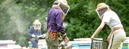 Two people wearing beekeeping protective gear hold a honeycomb up.
