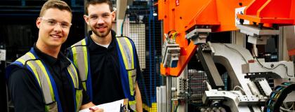 Two people wearing high visability vests smile at the camera, standing beside engineering automotive equipment.