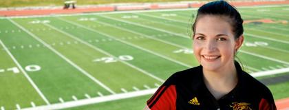 A person smiles at the camera wearing a red and black shirt, in front of a sports field.
