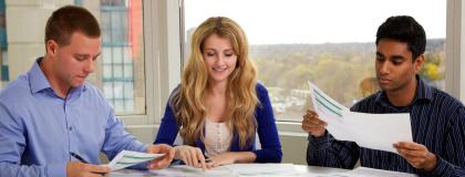 Three people sit at a desk looking at paper documents.