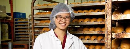 Person wearing a lab coat and a hair net smiles, standing beside bakers' racks of bread.