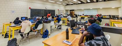 Students are in a physics lab being taught by a graduate teaching assistant, who is writing equations on a chalkboard while students observe.