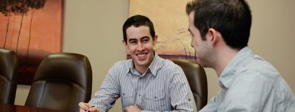 Two people talk to each other around a board room table.