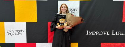 U of G Grad Jacquelyn Passarelli at her graduation standing in front of a U of G backdrop holding flowers.