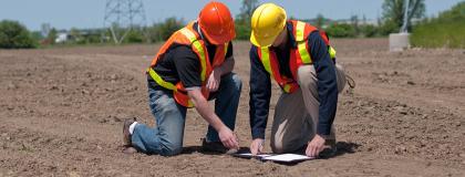 Two people wearing construction vests and hard hats kneel on dirt ground outside, looking at a paper.