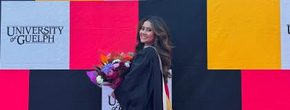 U of G grad in gown with flowers in front of the U of G logo
