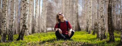 Women sitting cross legged on grass surrounded by birch trees. She is looking up at the sky